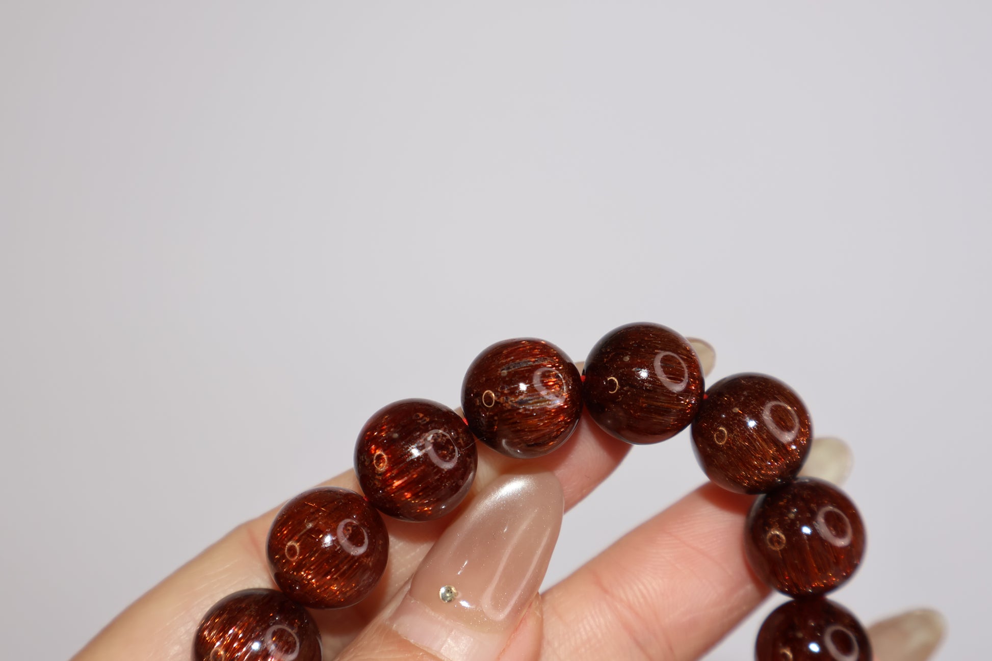 Close-up of Red Rutilated Quartz bracelet held between fingers, showing shimmering rutile inclusions and glossy bead surface with cat’s eye effect, natural gemstone jewelry.