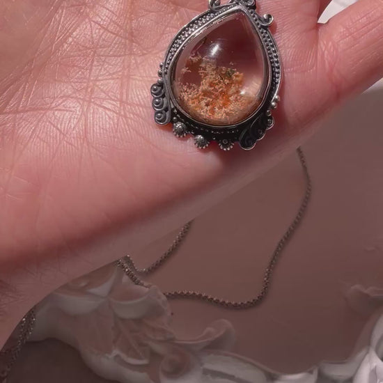 Video of a small teardrop Phantom Quartz pendant (17×22mm) with reddish-brown inclusions, rotating gently to reveal its clear crystal body and vintage S925 silver setting. The light catches its inner textures and reflections. Chain not included.