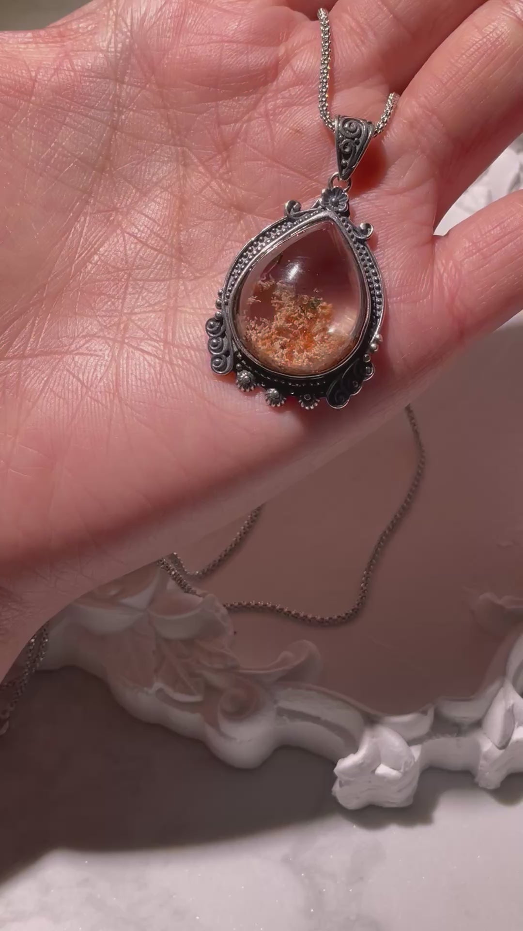 Video of a small teardrop Phantom Quartz pendant (17×22mm) with reddish-brown inclusions, rotating gently to reveal its clear crystal body and vintage S925 silver setting. The light catches its inner textures and reflections. Chain not included.