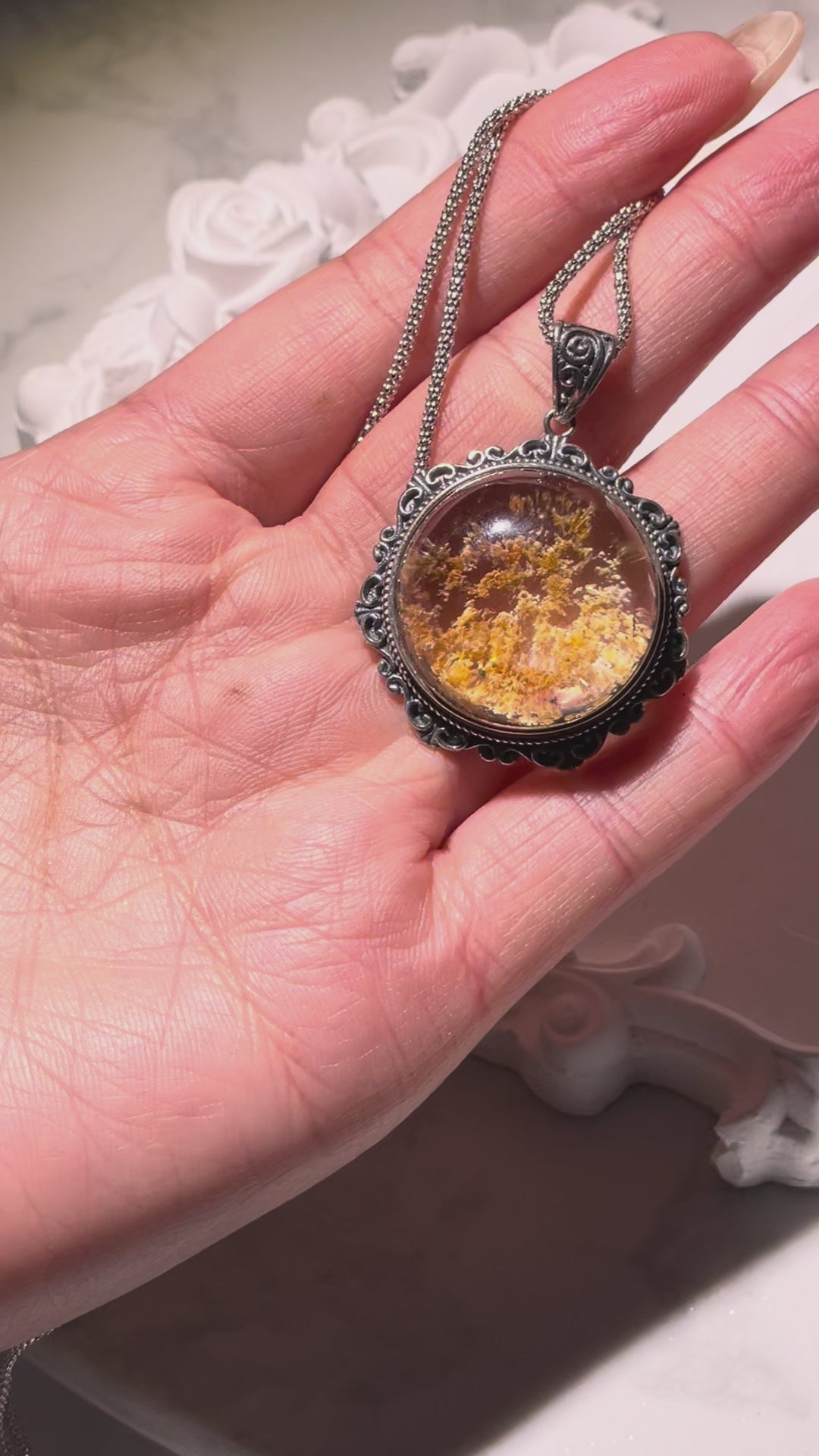 Video of a 24mm round Phantom Quartz pendant featuring golden-orange and dark mineral inclusions inside a domed crystal. The pendant is set in a detailed S925 vintage silver frame and rotates slowly under soft lighting to reveal its mystical internal patterns. Chain not included.