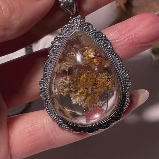 Video showcasing a large teardrop-shaped Phantom Quartz pendant (26×34mm) with red and golden inclusions and fine rutile threads. The crystal shimmers under light, revealing layered phantom formations inside a vintage S925 silver frame. Chain not included.