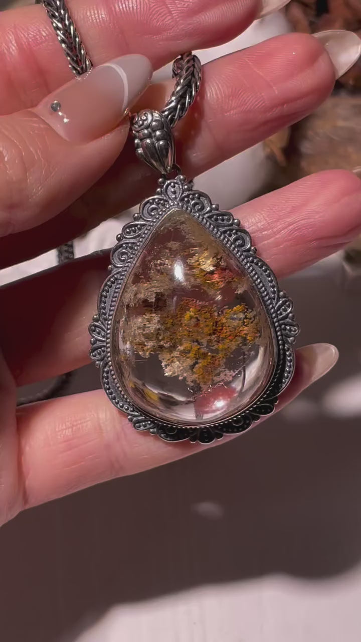 Video showcasing a large teardrop-shaped Phantom Quartz pendant (26×34mm) with red and golden inclusions and fine rutile threads. The crystal shimmers under light, revealing layered phantom formations inside a vintage S925 silver frame. Chain not included.