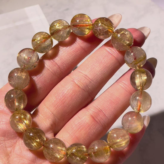 Video of an 11mm Golden Rutilated Quartz bracelet with red hair-like inclusions, rotating under natural light. Each bead sparkles with golden clarity and distinctive rutile threads, highlighting the rich golden and copper-red tones in motion.
